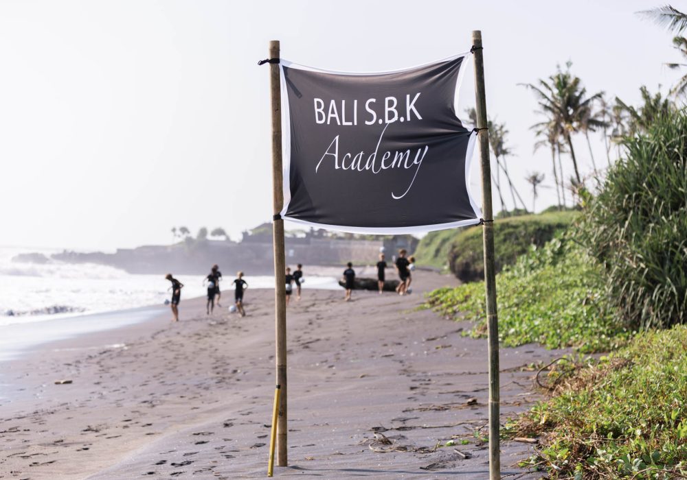 Team Bali SBK junior players train on beach, with football team's flag in foreground.